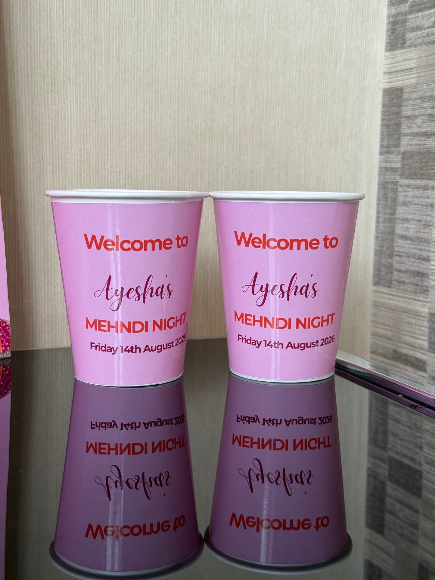 Two pink paper cups stacked on top of each other, printed with the text 'Welcome to Ayesha's Mehndi Night Friday 14th August 2026'. Paper cup colours are light pin and the writing it orange and pink. The papers cups are for custom events