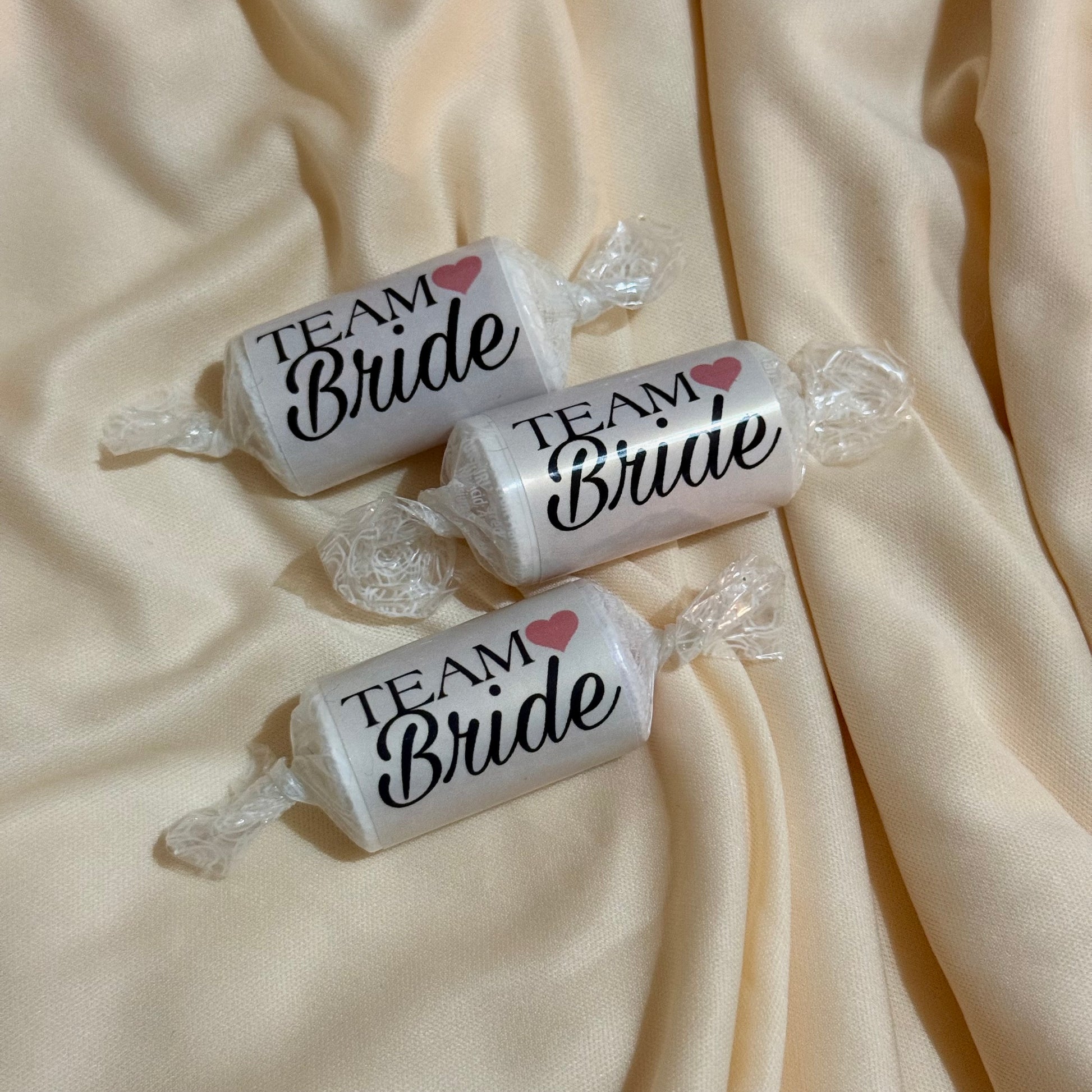Three wrapped candies on draped cream satin fabric, each printed with the words 'TEAM Bride' in black script with a small pink heart.