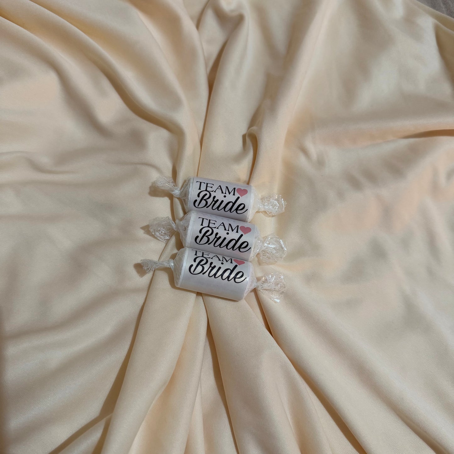 Three individually wrapped white candies with 'Team Bride' printed on them, arranged vertically on a draped cream satin fabric background.