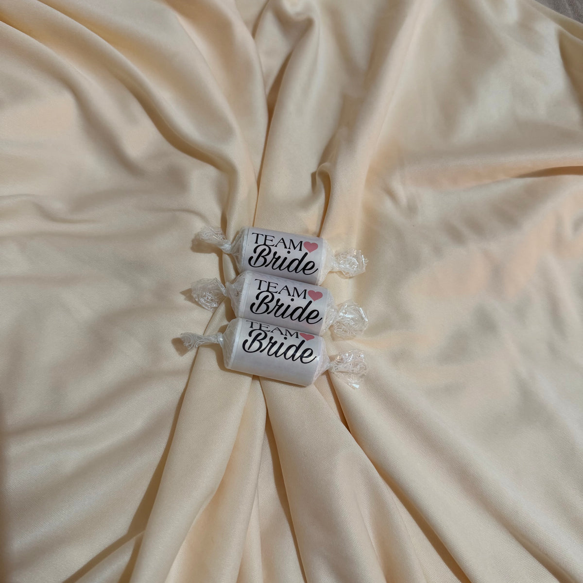 Three individually wrapped white candies with 'Team Bride' printed on them, arranged vertically on a draped cream satin fabric background.