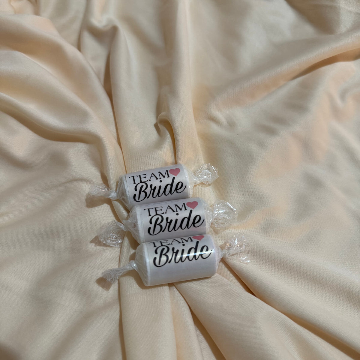 Three wrapped love heart favours  printed with 'TEAM Bride' in black script and a small pink heart, arranged on draped cream satin fabric.