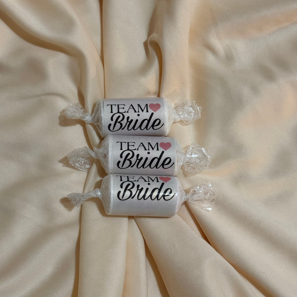 Three wrapped candies on a beige satin fabric background, each labeled 'TEAM Bride' with a small pink heart.