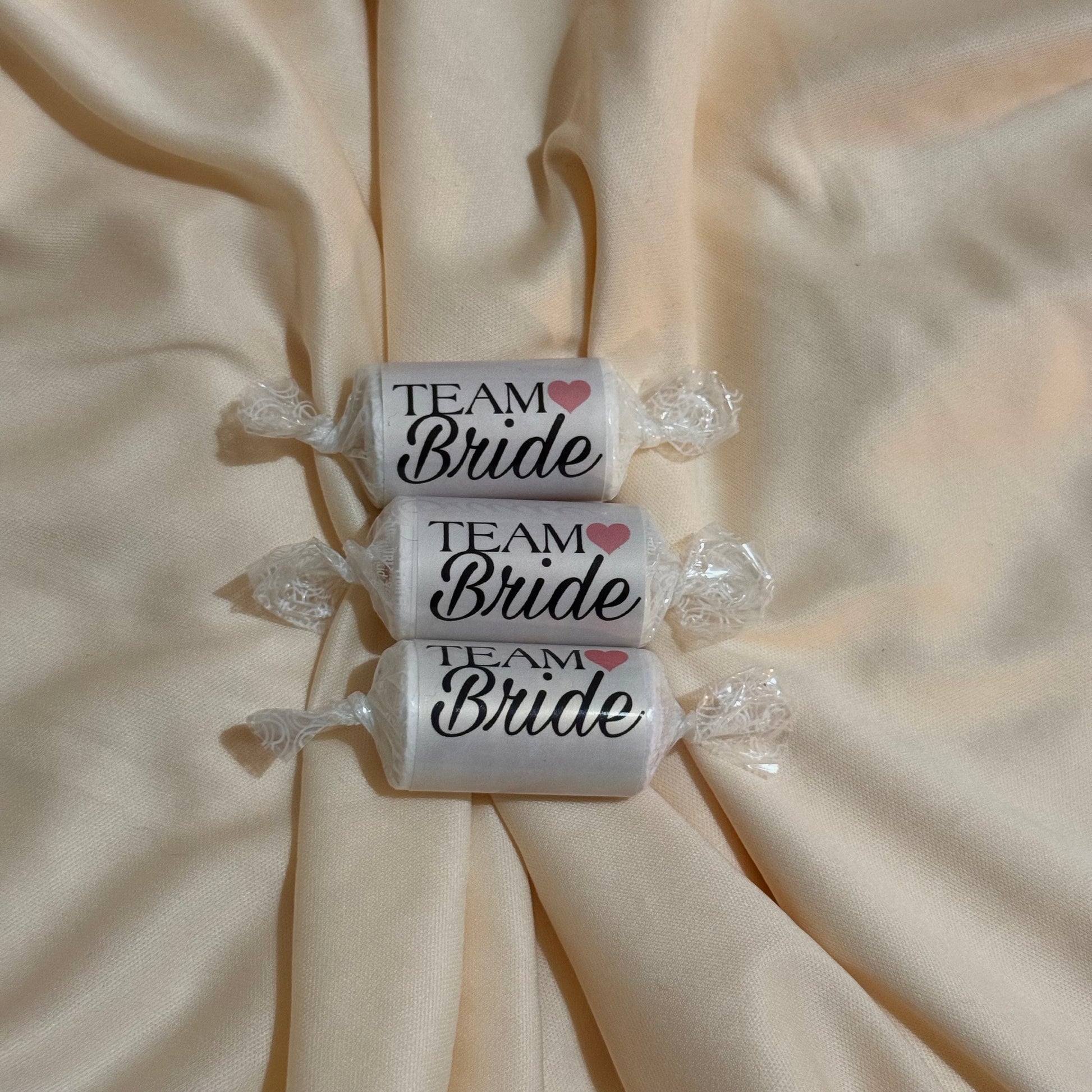 Three wrapped candies on a beige satin fabric background, each labeled 'TEAM Bride' with a small pink heart.