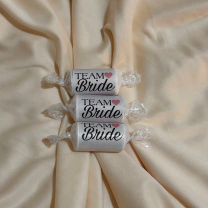 Three wrapped candies on a beige satin fabric background, each labeled 'TEAM Bride' with a small pink heart.